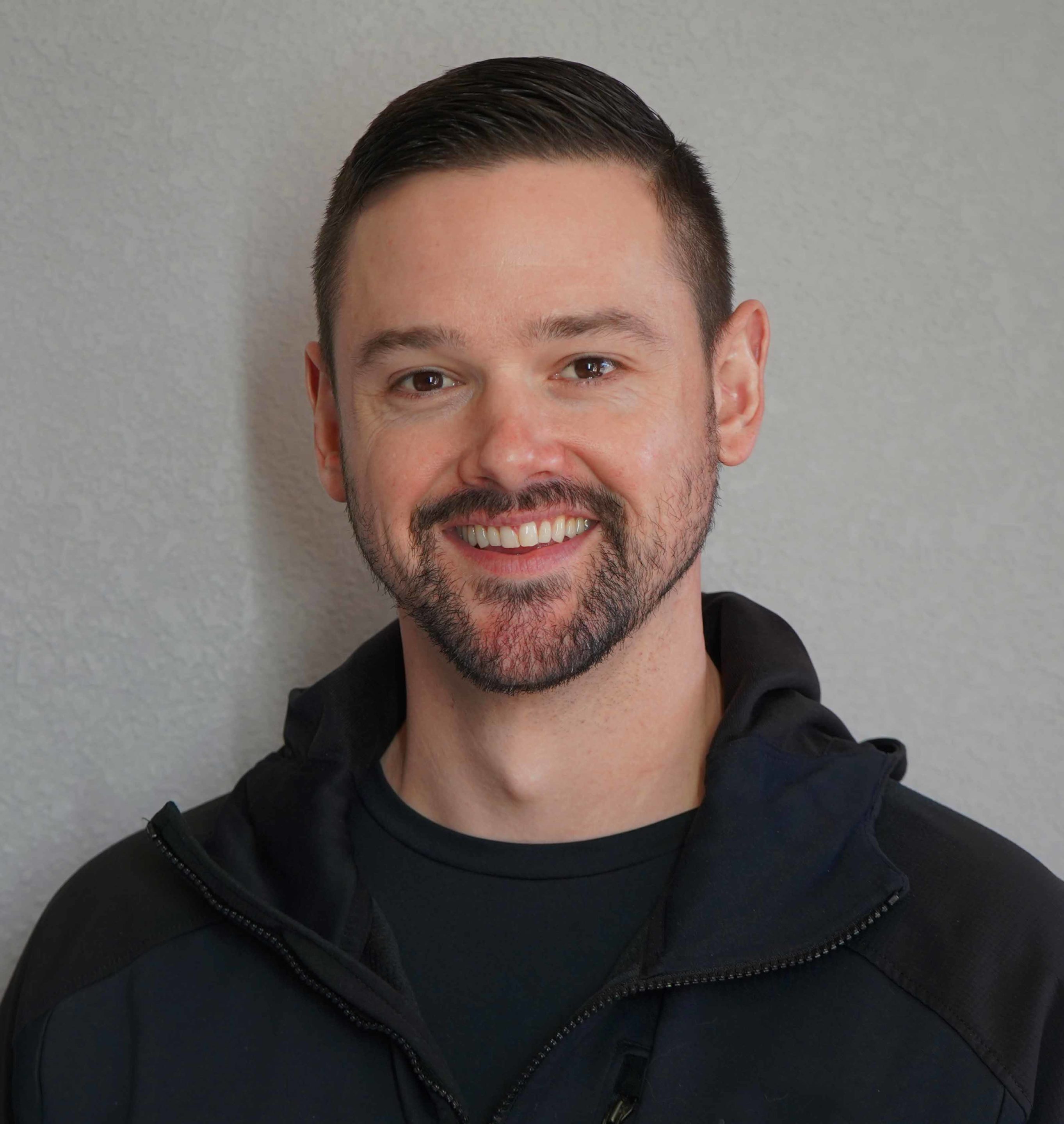 Man with short hair and beard, donning a black jacket, smiling confidently against a plain background, ready to start your personal training business.